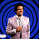 A game-show host in a shiny suit smiles at the camera.